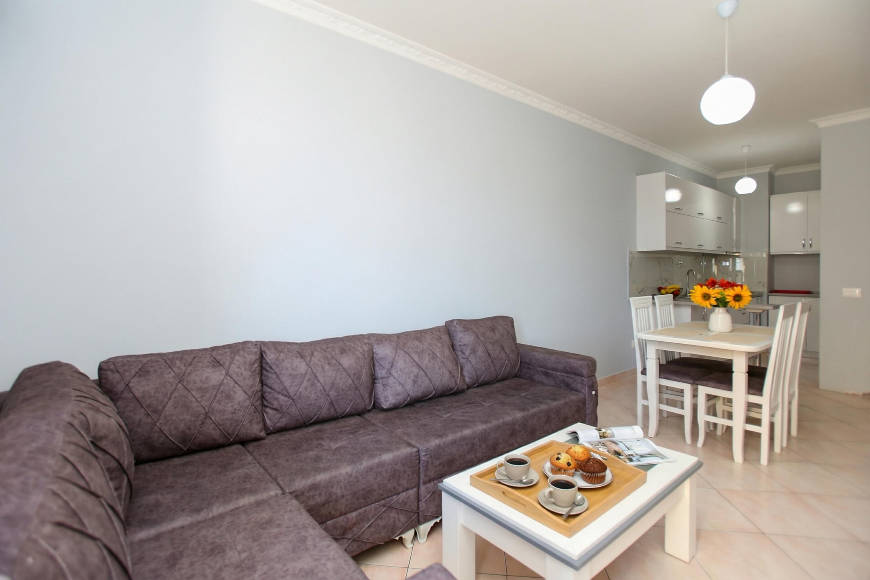 Un salon moderne avec un canapé d’angle, un coin repas avec une table et des chaises blanches, et une cuisine en arrière-plan.