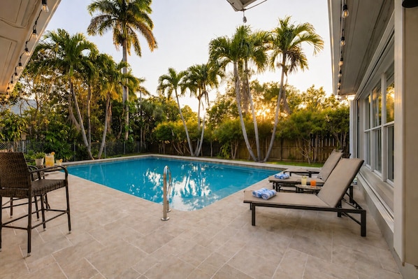 A swimming pool with lounge chairs, a table with drinks, and palm trees in the background.