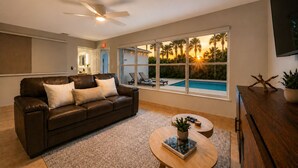 A living room with a leather sofa, a coffee table with a plant, a TV, and a large window with a pool view. - Luxury Pool Oasis • 5 Min to Beach • Modern Retreat (Hallandale Beach)