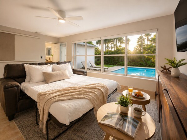A bedroom with a bed, a bedside table with a book and a cup, a view of a swimming pool, and a ceiling fan.