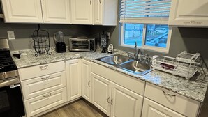 Una cocina con armarios blancos, encimera de granito, estufa, microondas, tostadora y una ventana con persianas.