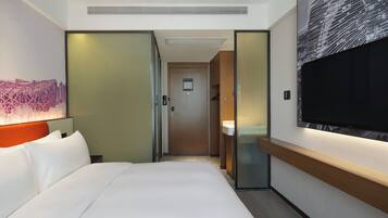Ein Hotelzimmer mit Bett, Fernseher, Bad und einem zur anderen Kammer führenden Gang.