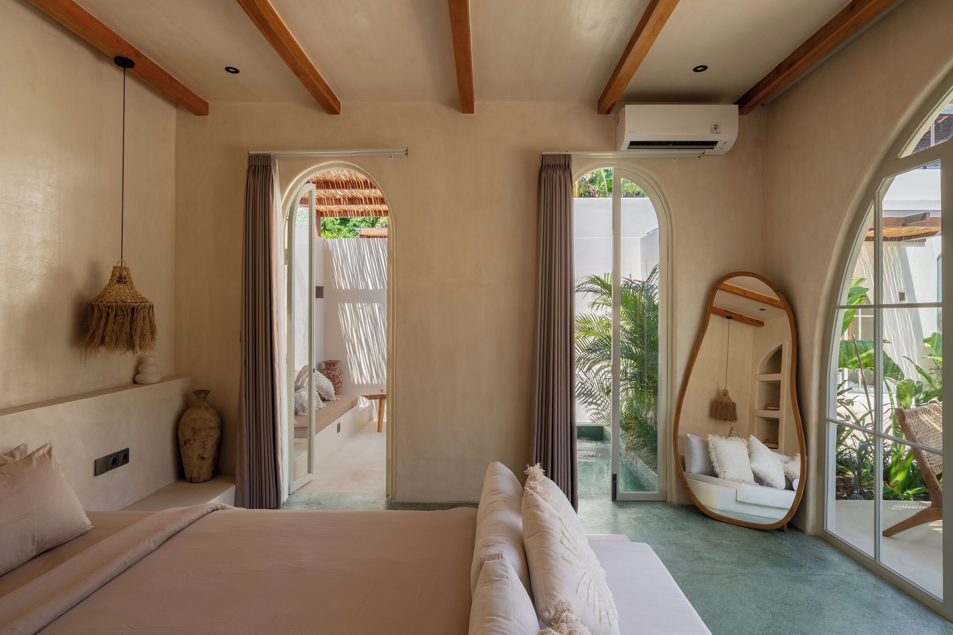 A bedroom with a large bed, a hanging light fixture, a mirror, and a view of the outdoors.