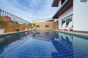 Una piscina con borde de azulejos, una pared de piedra y un edificio blanco con ventana y sillas.