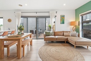 A modern living room with a wooden dining table, benches, a sectional sofa, and a large window. - Capel Haven House by Ready Set Stay (Capel Sound)