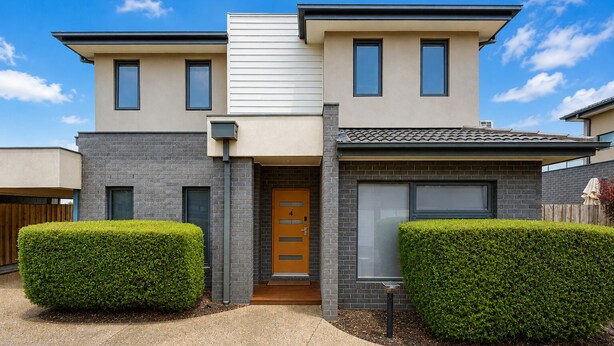 Una casa moderna de dos pisos con techo plano, una puerta principal con un cartel de número y dos setos bien cuidados.