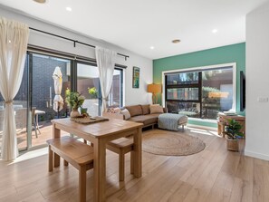 A modern living room with a wooden dining table, a sofa, and a large window. - Capel Haven House by Ready Set Stay (Capel Sound)