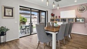 A dining area with a wooden table and grey chairs, a sliding glass door leading to a patio, and a wall clock. - Garden Retreat 4-Bed House in Tarneit (Tarneit)