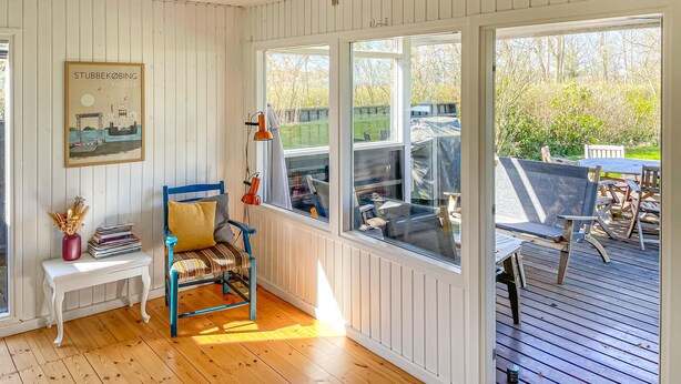 Een kamer met een houten vloer, een blauwe stoel, een witte bank, een ingelijst schilderij en uitzicht op een terras met stoelen en een tafel.