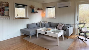En moderne stue med en grå hjørnesofa, et glas sofabord og en sort gyngestol. To vinduer med hvide skodder, en hvid væg med indbygget reol og en aircondition-enhed monteret på væggen.