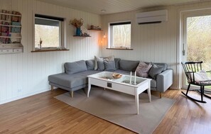 A modern living room with a grey sectional sofa, a glass coffee table, and a black rocking chair. There are two windows with white shutters, a white wall with a built-in shelf, and an air conditioning unit mounted on the wall. - Amazing home in Dannemare with WiFi (Dannemare)