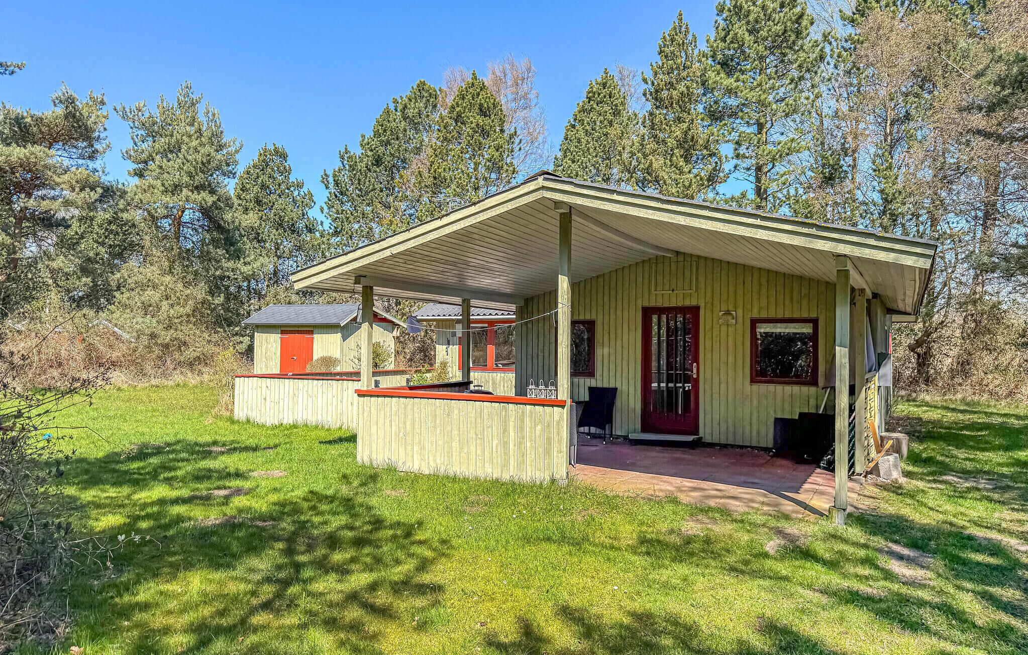 Een klein houten huisje met een veranda, een rode deur en een kleine schuur, gelegen in een bebost gebied.