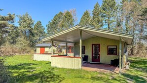 Een klein houten huisje met een veranda, een rode deur en een kleine schuur, gelegen in een bebost gebied.