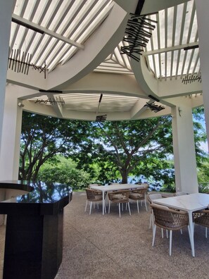A covered outdoor seating area with tables and chairs, a black counter, and a view of trees and sky. - Cozy apartment w/parking/pool/gym/viewsA (bucerias)