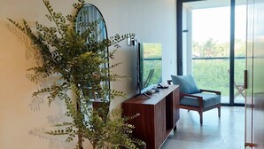 Een moderne woonkamer met een grote potplant, een houten console, een televisie en uitzicht door een glazen deur op een buitenruimte.