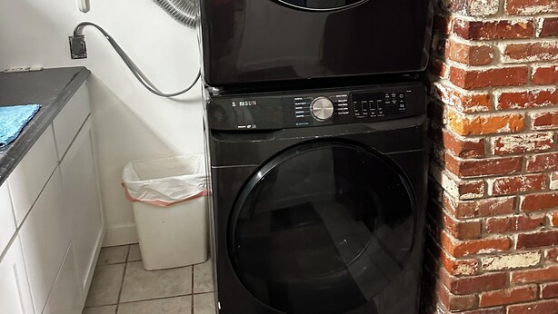 Une buanderie équipée d’une pile de lave-linge et de sèche-linge, d’une petite étagère contenant des produits de nettoyage et d’un sol carrelé.