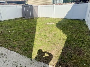 A backyard with a white fence, a shed, and a shadow of a person. - Luxe 2BR Apt. w/ backyard Near Airport + Hospital (Newark)