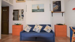 Ein Wohnzimmer mit einem blauen Sofa, orangefarbenen Beistelltischen, einer Tür und einem an der Wand hängenden Kunstwerk.
