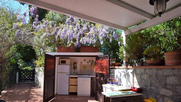 Eine kleine Küche mit Kühlschrank, Mikrowelle und Spüle, unter einer Pergola mit violetten Blüten.