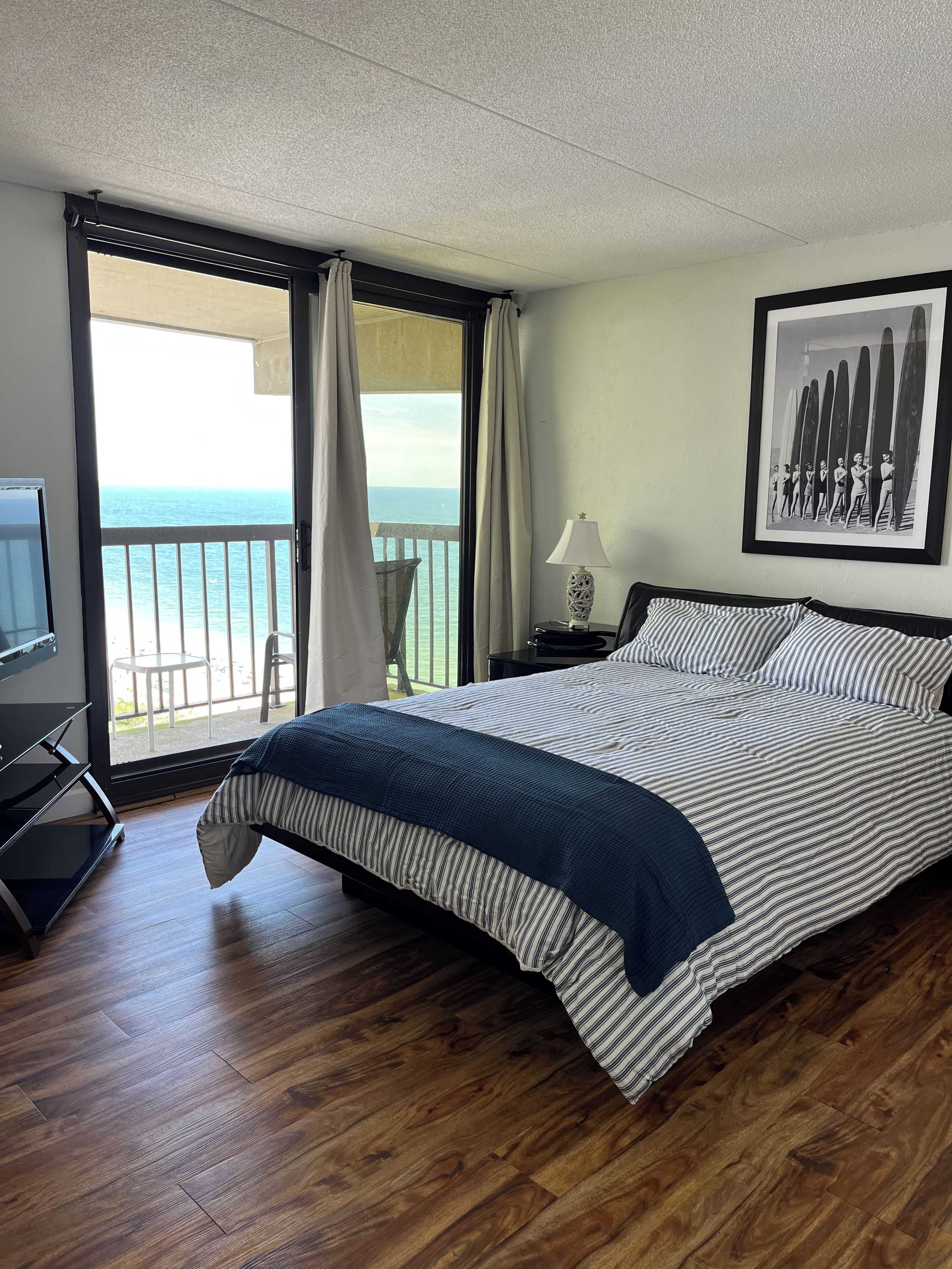 A hotel room with a bed, a TV, a balcony with a view of the sea, and a framed picture on the wall.