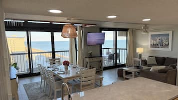 Zona saló àmplia amb un balcó que domina la platja, taula de dinar per a quatre i televisió de pantalla plana penjada a la paret.