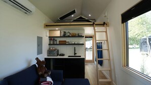 A compact kitchen with a microwave, toaster, and sink. A wooden ladder is against the wall, and a window reveals a view of trees.
