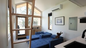 A modern room with a loft bed, a blue sofa, a small stool, and a window with a view of trees.