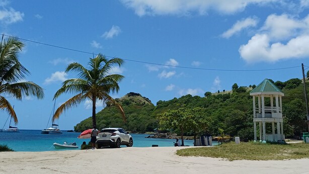 A beach with a white car parked on the sand, palm trees, a small gazebo, and boats in the water.