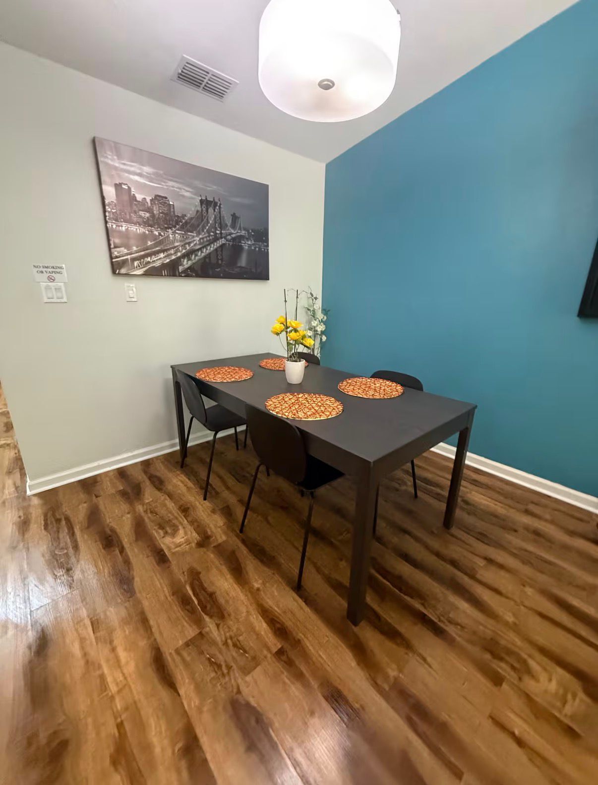 Uma sala de jantar com mesa posta para quatro pessoas, uma pintura de paisagem urbana e um vaso com flores.