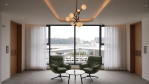 Une chambre d’hôtel moderne dotée d’une grande fenêtre, de deux fauteuils en cuir vert, d’une petite table ronde et d’un luminaire au plafond au design original.