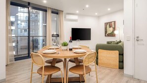 A modern living room with a round dining table, four chairs, a green sofa, a flat-screen TV, and a window with curtains. - LaneStay - 2BR in Southbank w Large Living and Gym (Southbank)