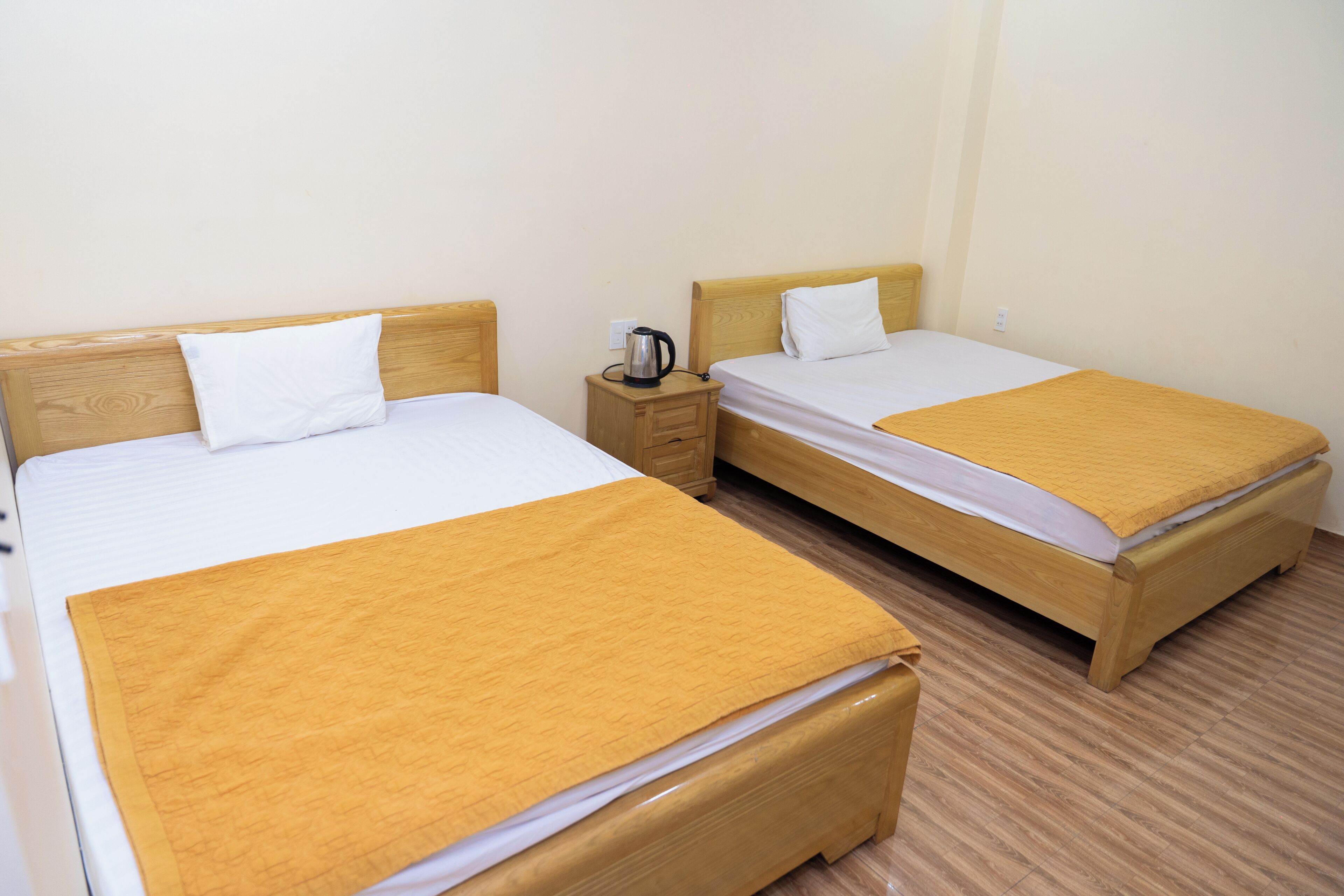 Two single beds with wooden headboards, a nightstand with a kettle, and a plain wall.
