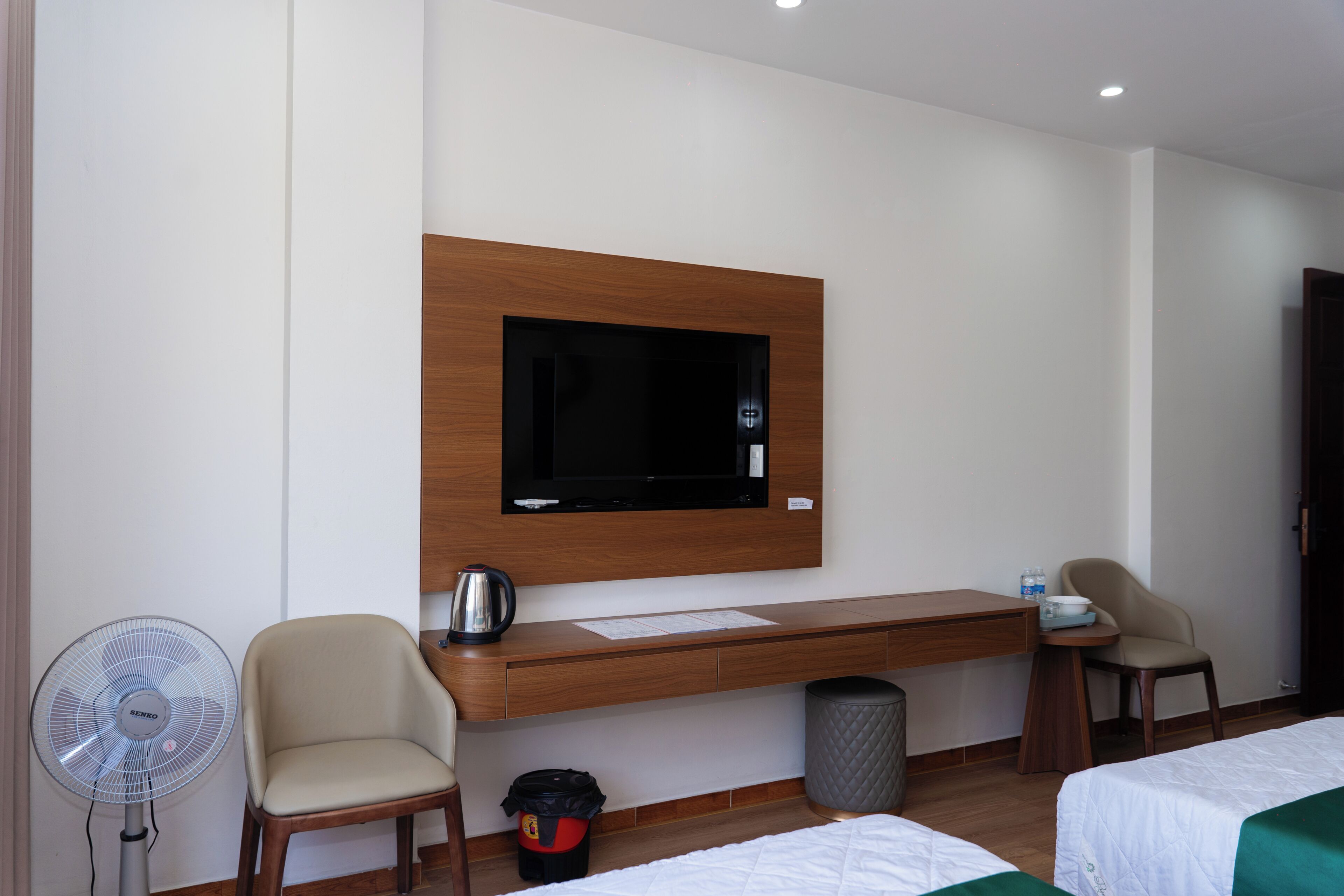 A hotel room with a wooden desk, two chairs, a television mounted on the wall, a kettle, a trash bin, and two beds with green bedspreads.