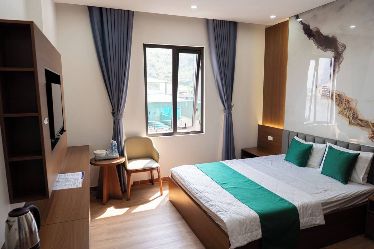 Quarto de hotel com cama, cadeira, mesa pequena, chaleira e janela com cortinas.