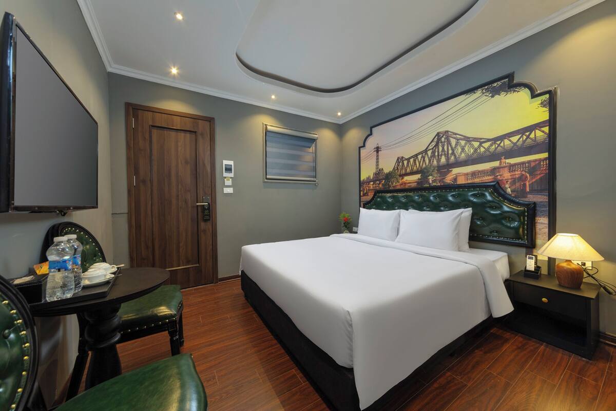A hotel room with a bed, a desk with a chair, a TV, a painting, and a nightstand with a lamp.