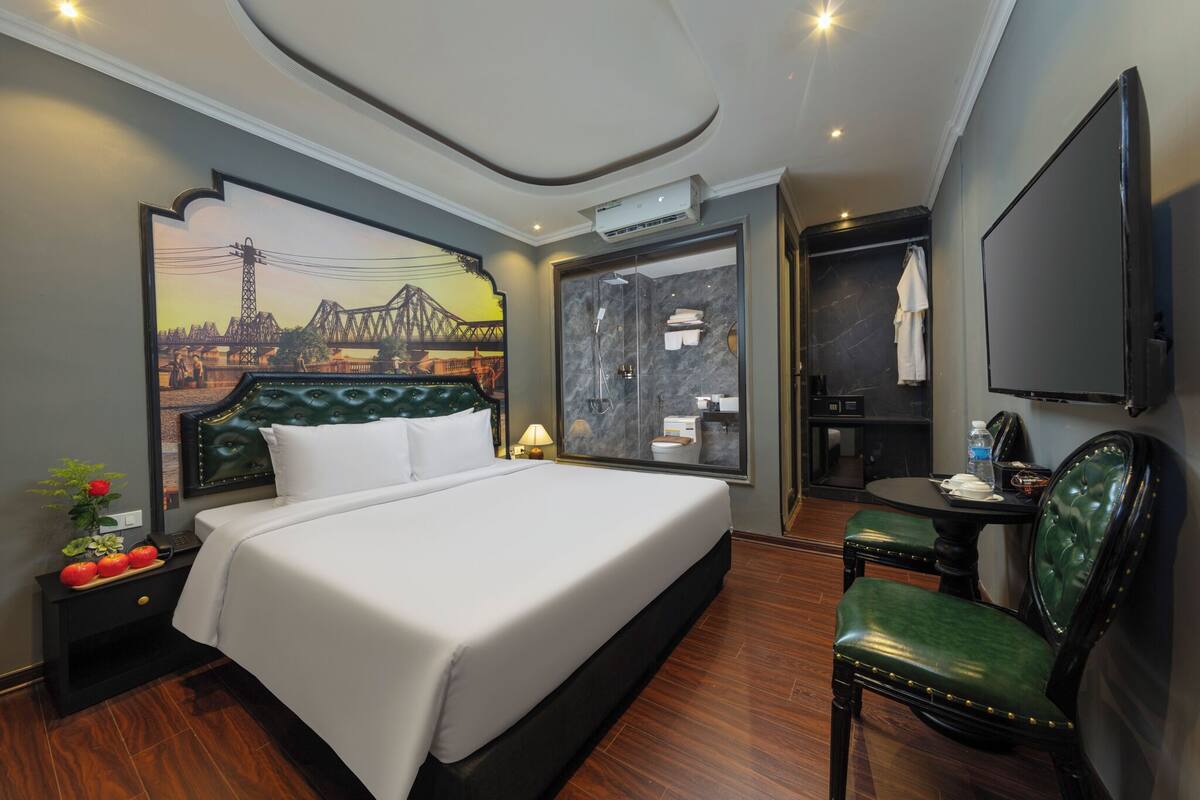A hotel room with a bed, a desk with a chair, a TV, a shower area, and a wall with a cityscape mural.