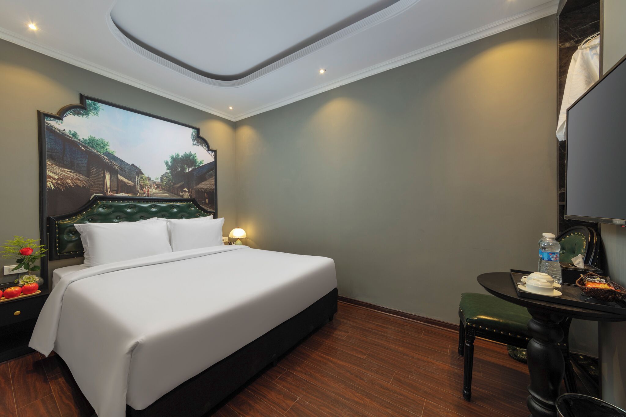 A hotel room with a large bed, a small table with a chair, a painting on the wall, and a ceiling light.