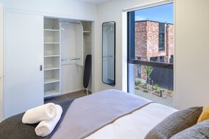 A bedroom with a bed, a large mirror, a wardrobe, and a view of a brick building through a window. - City Comfort with Carpark - Walk to Hagley Park (Christchurch)