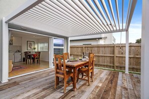 A wooden deck with a table and chairs, a pergola, and a view of a backyard. - Aiturangi Retreat Whangamata (Whangamata)