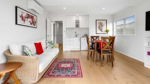 A modern living room with a sofa, dining table, and chairs, a kitchen area, and a large window with blinds. - Aiturangi Retreat Whangamata (Whangamata)