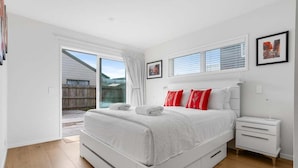 A bedroom with a bed, a nightstand, a window with blinds, and a door leading outside. - Aiturangi Retreat Whangamata (Whangamata)