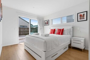 A bedroom with a bed, a nightstand, a window with blinds, and a door leading outside. - Aiturangi Retreat Whangamata (Whangamata)