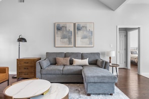 Een moderne woonkamer met een grijze modulaire bank, een houten bijzettafeltje, een vloerlamp en abstracte kunst aan de muur.
