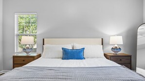 Uma cama bem arrumada com colcha listrada em branco e cinza, um travesseiro azul e duas luminárias nas mesas de cabeceira de madeira.