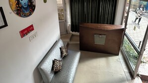 A reception area with a sofa, a reception desk, a wall-mounted artwork, a sign with operating hours, and a window with a view of the outside. - Hotel Devidyar (Manali)
