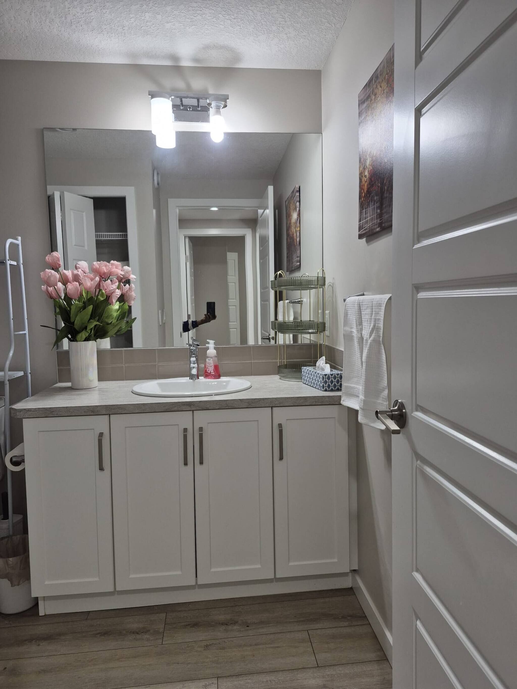 A bathroom with a white vanity, a mirror, a sink, and a vase of pink flowers.