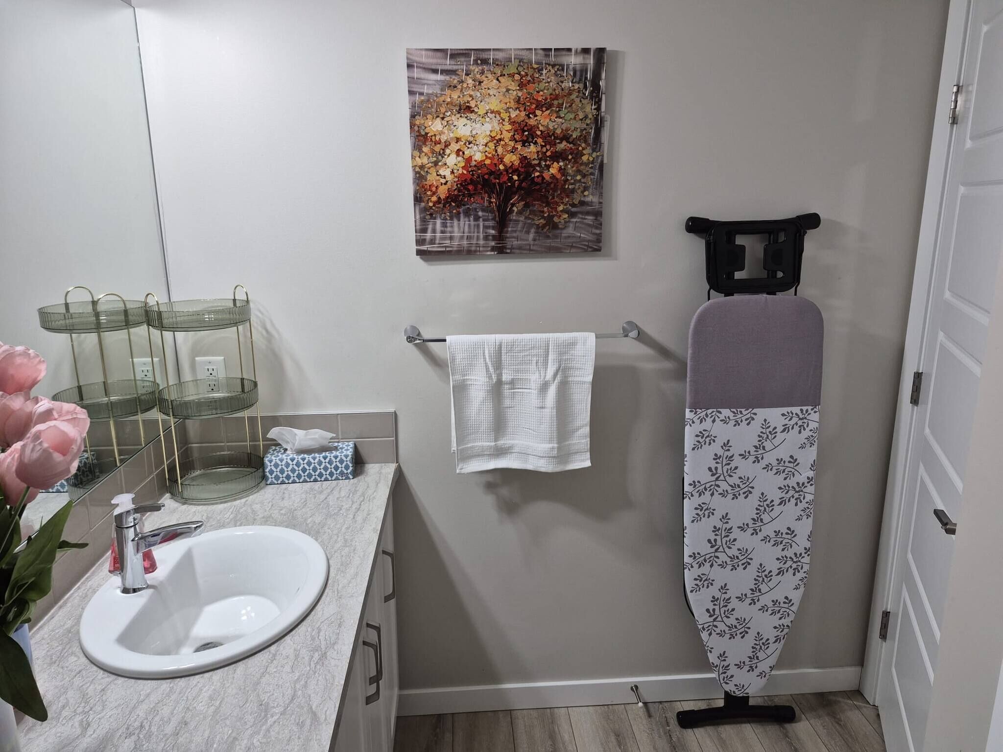 Ein Badezimmer mit einem weißen Waschbecken, einem Spiegel, einem Handtuchhalter, einem Baumgemälde und einem schwarzen Bügelbrett.