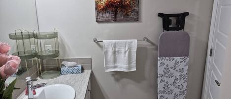Ein Badezimmer mit einem weißen Waschbecken, einem Spiegel, einem Handtuchhalter, einem Baumgemälde und einem schwarzen Bügelbrett.