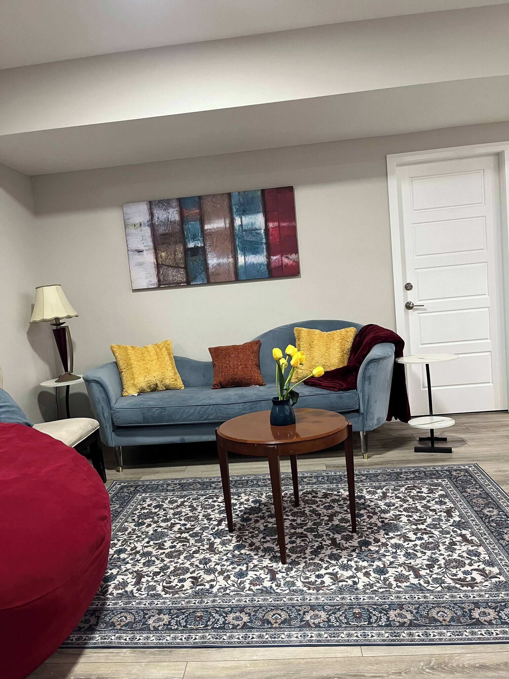 Ein Wohnzimmer mit einem blauen Sofa, einem roten Sessel, einem kleinen Tisch mit einem Blumenstrauß aus gelben Blumen, einer Lampe und einer Tür.