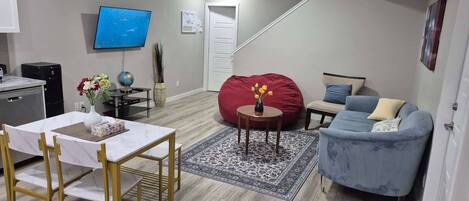 A modern living room with a dining area, a flat-screen TV, a red bean bag, a blue sofa, and a staircase leading upstairs.